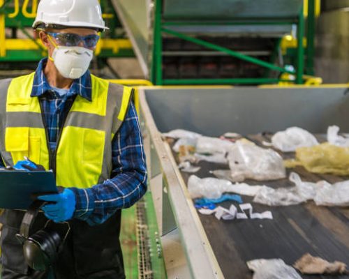 istockphoto-1567731426-612×612 Waste Management Sorting Facility Caucasian Worker Preparing Documentation. Wearing Air Pollution Mask, Eyes Safety Glasses and a Hard Hat.